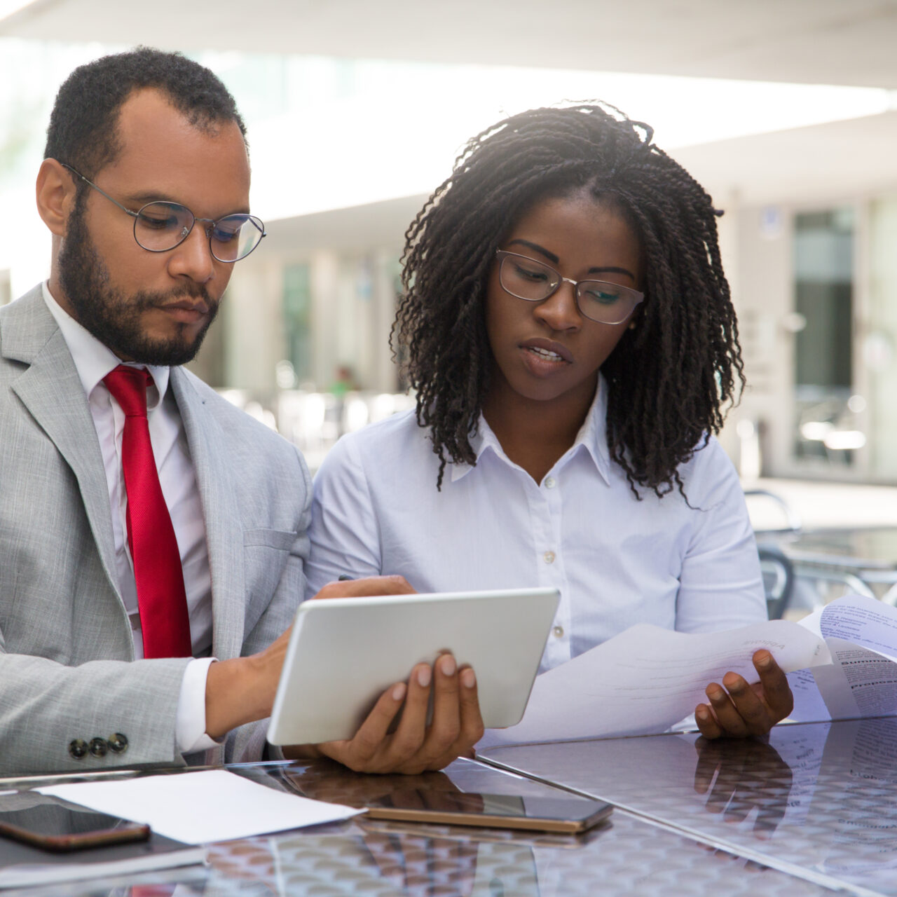 Focused coworkers checking reports on papers and on tablet. Business man and woman sitting in coffee shop, showing tablet and smartphone screens to each other. Teamwork concept