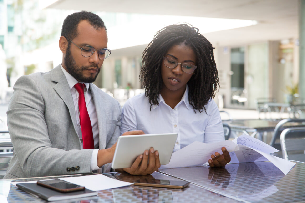 Focused coworkers checking reports on papers and on tablet. Business man and woman sitting in coffee shop, showing tablet and smartphone screens to each other. Teamwork concept